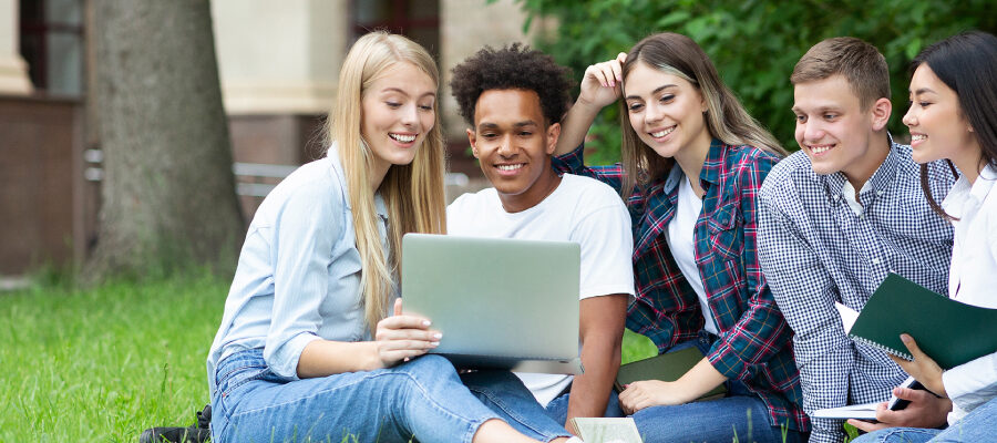 A diverse group of five students sit on grass, smiling and engaging with a laptop. The setting is outdoors, creating a relaxed and collaborative atmosphere.