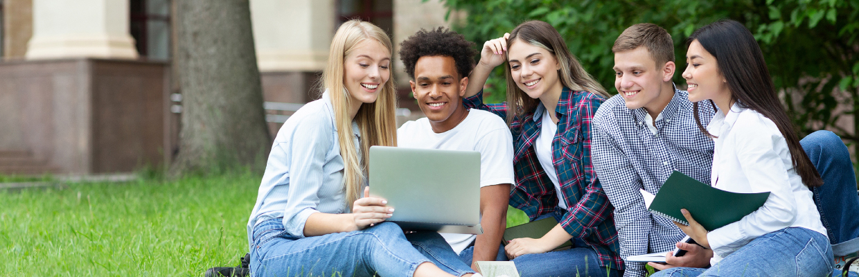 A diverse group of five students sit on grass, smiling and engaging with a laptop. The setting is outdoors, creating a relaxed and collaborative atmosphere.