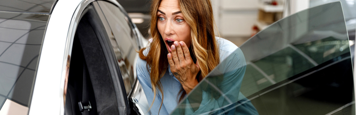 A woman with long brown hair wears a light blue shirt, looking surprised and excited by a new car. Her hand covers her mouth, and the car door is open.