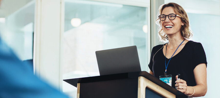 A smiling woman stands at a lectern, delivering a presentation. She's wearing glasses and a black top, suggesting a professional and confident atmosphere.