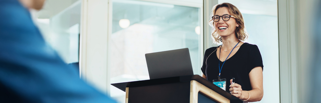 A smiling woman stands at a lectern, delivering a presentation. She's wearing glasses and a black top, suggesting a professional and confident atmosphere.