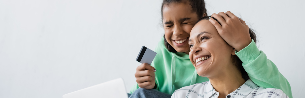 A joyful mother and daughter sit together, smiling with excitement. The daughter holds a credit card, and a laptop is visible, suggesting online shopping.