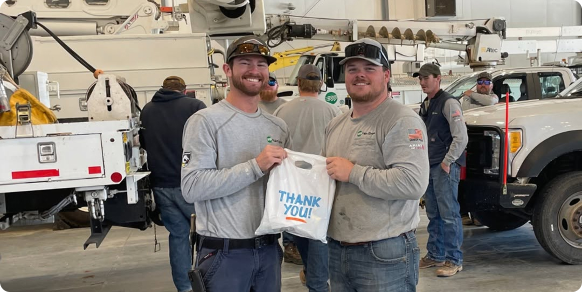Two smiling linemen holding a white bag with "THANK YOU!" printed on it, in a workshop with trucks